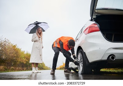 Male auto mechanic unscrewing lug nuts on car wheel while elegant woman holding umbrella. Young man in vest repairing woman automobile on the road. Concept of emergency road service. - Powered by Shutterstock