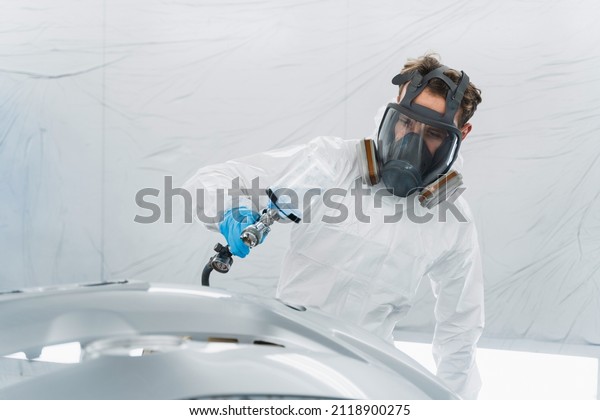 Male auto painter in protective mask\
and suit painting automobile car bumper in chamber\
