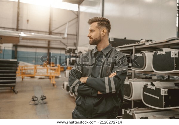Male\
aviation maintenance technician keeping arms crossed and looking\
away while standing in aircraft repair\
station