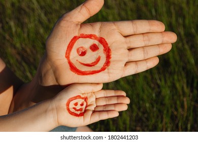 Male And Baby's Palm With Paint Red Face And Smile On Green Grass Background