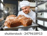 Male baker holding a tray of baked breads in bakery shop.