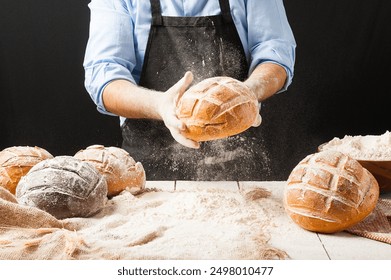 Male baker wearing black apron holding a freshly baked rustic bread splashing flour in front of a white wooden table filled with different kinds of loaves and jutte fabric    - Powered by Shutterstock