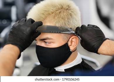 A Male Barber Gives A Haircut To A Client Who Is Wearing A Protective Mask. A Barber In Nitrile Gloves Uses Scissors And A Comb.