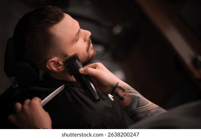 A male barber with tattooed arms is trimming a client's beard using electric clippers. The client is reclined in a chair, eyes closed, enjoying a professional grooming session in a modern barbershop.
