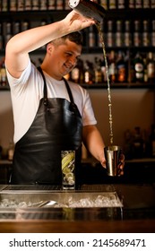 Male Bartender In Black Apron Skillfully Pours A Cocktail From One Shaker Cup To Another