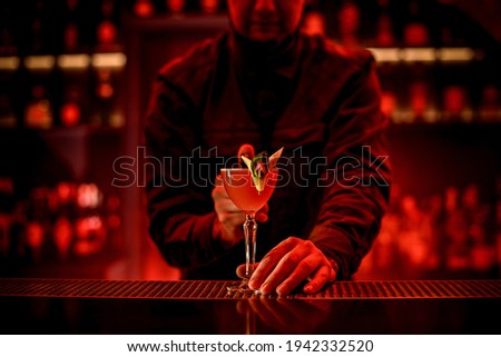male bartender holds with his hand beautiful glass with alcoholic cocktail on bar counter. Red illumination.