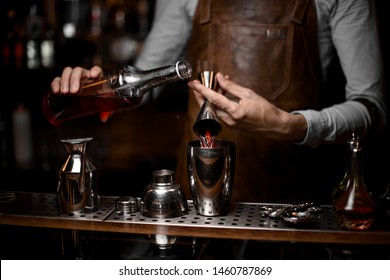 Male bartender in leather apron pouring alcohol from jigger to shaker