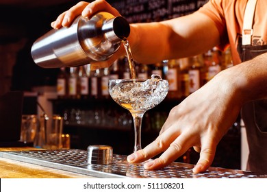 Male Bartender Is Making Cocktail Pouring Alcohol From Shaker To Glass At Bar Background. Barman At Work.