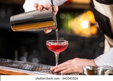 Male Bartender Is Making Cocktail Pouring Alchohol From Shaker To Glass At Bar Background.