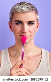 Male Beauty Blogger Holding Make-up Brush Near Mouth And Looking At Camera Isolated On Purple Background, Smiling