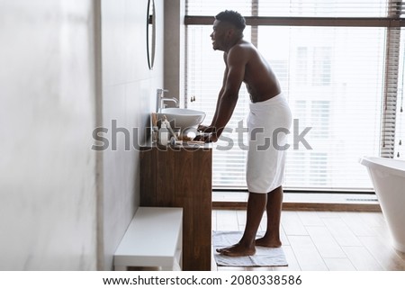 Male Beauty And Bodycare Routine. Happy African American Man Standing Wrapped In Towel After Shower Looking In Mirror, Smiling To His Reflection In Modern Bathroom At Home. Full Length, Side View