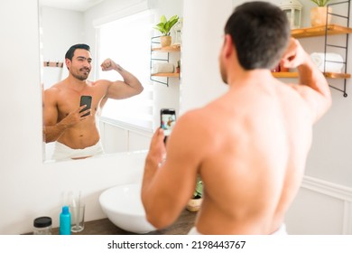 Male Beauty Concept. Muscular Happy Man Doing A Bicep Curl While Taking A Selfie In The Bathroom Mirror With His Smartphone