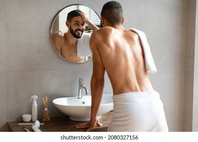 Male Beauty And Haircare Routine. Shirtless Handsome Man Touching Hair Looking In The Mirror After Shower, Smiling To His Reflection Standing In Modern Bathroom At Home, View Over The Shoulder