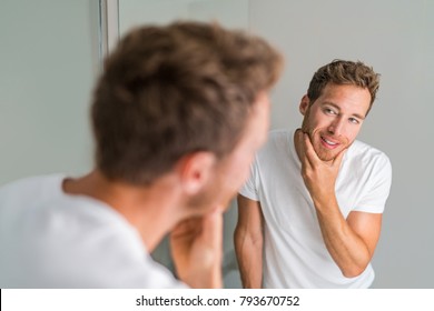 Male Beauty Young Man Touching Beard And Face Looking In Mirror - Healthy Skin. Skincare In Home Bathroom. After Shave Men Lifestyle Shaving Concept.