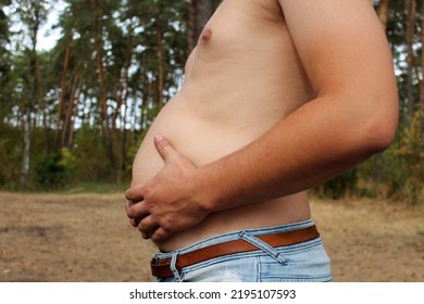 
Male Beer Belly, A Man Suffering From Overweight, Going On A Diet, A Fat Male Belly, Body Positivity, A Man Hugs A Big Belly But Against The Backdrop Of A Pine Forest, The Belly Hangs Out Of Pants