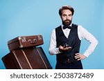 Male bellboy with baggage and bell used to ring for service or help at hotel reception in hospitality industry. Young employee working as doorkeeper or porter handling suitcases.