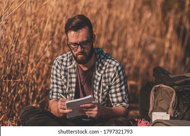 Male Biologist Sitting On Wooden Lake Docks On A Sunny Spring Day, Taking Notes While On Field Work Trip