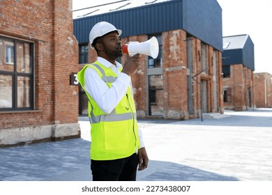 Male boss shouting in megaphone with aggressive facial expression while standing outdoors. Wicked african american architect in helmet giving orders to workers at construction site - Powered by Shutterstock