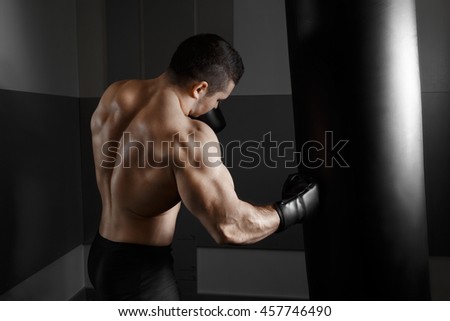 Male boxer training with punching bag in dark sports hall. Young boxer training on punching bag. Male boxer as exercise for the big fight. Boxer hits punching bag. Young masculine male athlete.
