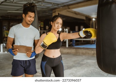 a male boxing coach standing next to the female boxer bring the clipboard while training her to punch the heavy bag at the boxing studio - Powered by Shutterstock