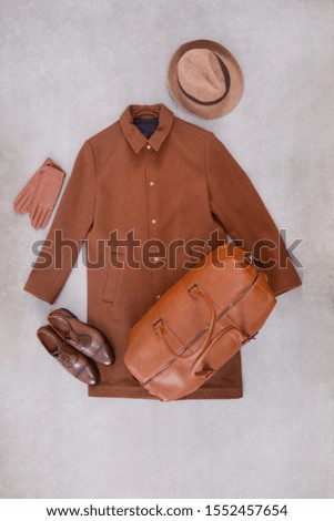 Male brown long coat with leather brown shoes with large leather handbag ,hat ,gloves on gray background 


