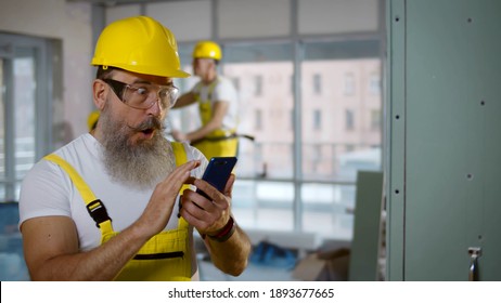 Male Builder Browsing Mobile Phone During Break And Doing Winner Gesture Getting Good News. Aged Excited Foreman In Hardhat And Uniform Using Smartphone Raising Fist Celebrating Success