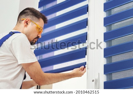 Male builder carpenter pulling down a blue jalousie on a window. Repairing and inspection fabric curtains. Duo system day and night blind. Interior decor element
