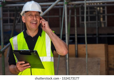 Male builder foreman, worker, contractor or architect on construction site holding black clipboard and talking on his cell phone