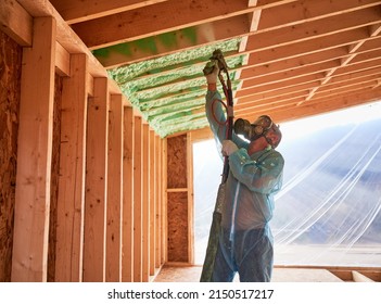 Male Builder Insulating Wooden Frame House. Man Worker Spraying Polyurethane Foam Inside Of Future Cottage, Using Plural Component Gun. Construction And Insulation Concept.