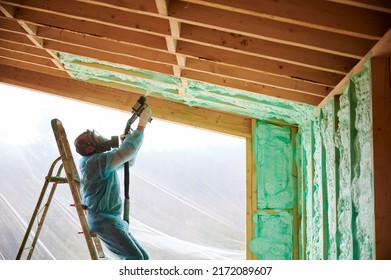 Male Builder Insulating Wooden Frame House. Man Worker Spraying Polyurethane Foam Inside Of Future Cottage, Using Plural Component Gun. Construction And Insulation Concept.