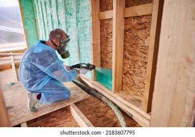 Male Builder Insulating Wooden Frame House. Man Worker Spraying Polyurethane Foam Inside Of Future Cottage, Using Plural Component Gun. Construction And Insulation Concept.