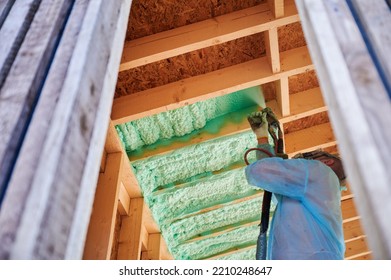 Male Builder Insulating Wooden Frame House. Man Worker Spraying Polyurethane Foam Inside Of Future Cottage, Using Plural Component Gun. Construction And Insulation Concept.