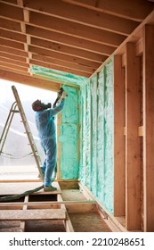 Male Builder Insulating Wooden Frame House. Man Worker Spraying Polyurethane Foam Inside Of Future Cottage, Using Plural Component Gun. Construction And Insulation Concept.