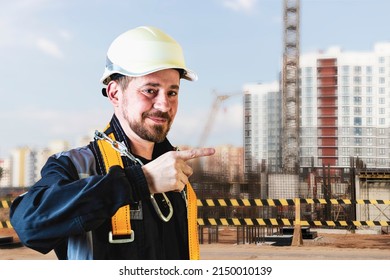 A Male Builder In A White Hard Hat Against A Blurred Background Of A Construction Site With A Blue Sky. Positive Civil Engineer With A Beard