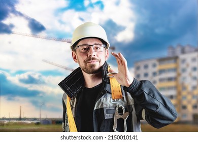 A Male Builder In A White Hard Hat Against A Blurred Background Of A Construction Site With A Blue Sky. Positive Civil Engineer With A Beard