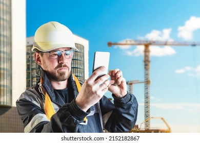 A Male Builder In A White Hard Hat Against A Blurred Background Of A Construction Site With A Blue Sky. Positive Civil Engineer With A Beard