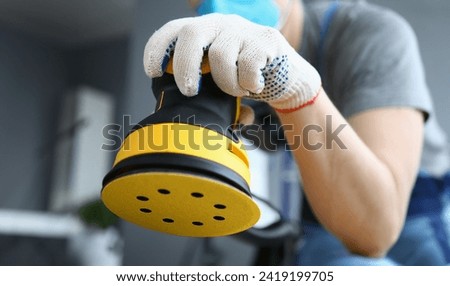 Male builder in work gloves holds sander machine. Preliminary cleaning and polishing surfaces. Carpenter works with tree with sander machine. Man makes home repairs during quarantine