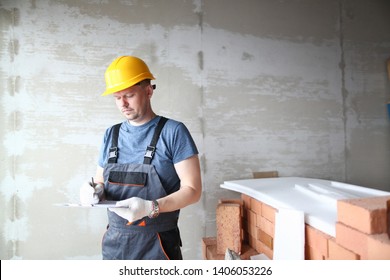 Male Builder In Yellow Hard Hat Holds Clipboard On Background Of An Apartment Under Repair Portrait. Estimated Property Value Concept.