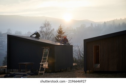 Male Builders Installing Black Corrugated Iron Sheet Used As Facade Of Future Cottage In The Evening. Men Workers Building Wooden Frame House. Carpentry And Construction Concept.