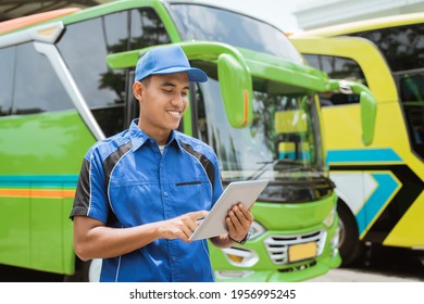 A Male Bus Crew Member In Uniform And A Hat Smiles While Using A Digital Tablet Against The Backdrop Of The Bus Fleet