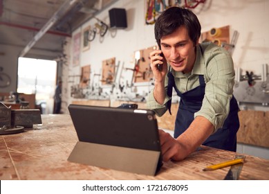 Male Business Owner In Workshop Using Digital Tablet And Making Call On Mobile Phone