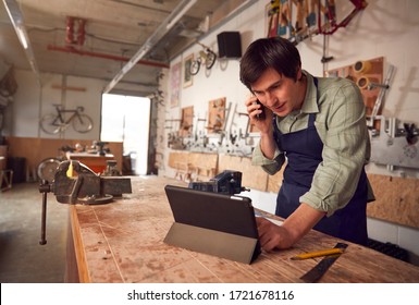 Male Business Owner In Workshop Using Digital Tablet And Making Call On Mobile Phone