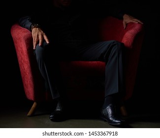 Male Businessman With A Clock On His Hand, In A Black Suit Sitting In A Red Chair, Black Background, No Faces Visible, Studio Shooting