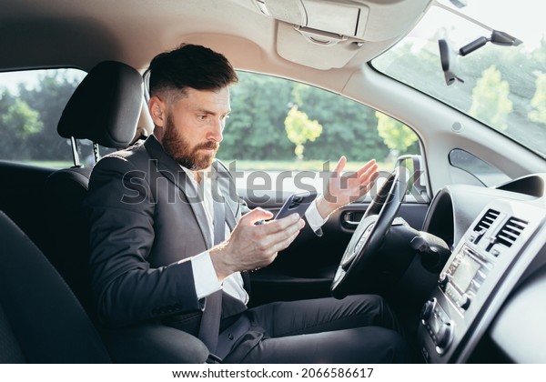 Male businessman\
sitting behind the wheel of a car, frustrated can not understand\
the steering and instructions funky car, reads additional\
information from the phone