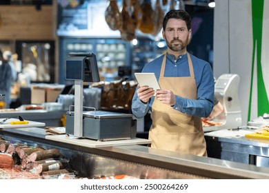 Male butcher in apron using tablet behind counter at deli shop. Display of various meats and sausages visible. Digital transformation in traditional retail setting - Powered by Shutterstock