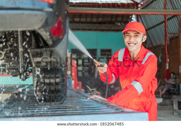Male car cleaner smiles\
wearing red uniform and hat while spraying water on the car in the\
car salon