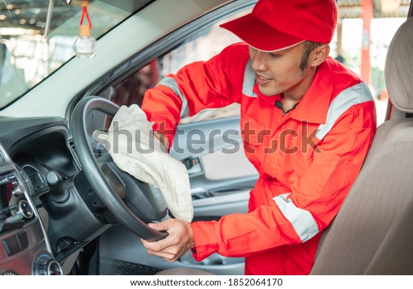 Male car cleaner wearing red uniform wipes the\
steering wheel in car\
salon