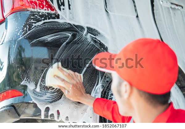 Male car cleaner wearing red\
uniform and hat washes car bumper with foaming sponge in car\
salon