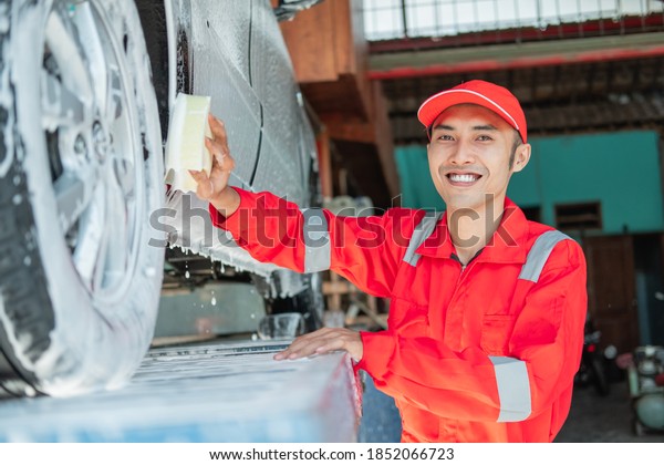 Male car cleaner wears\
red uniform and smiling hat while washing the bottom of the car in\
the car salon