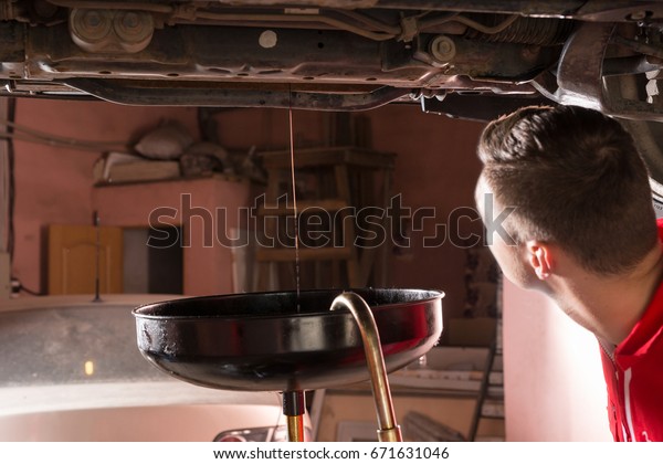 Male car mechanic in uniform\
watching how oil flows out and changing motor oil in automobile\
engine at maintenance repair service station in a car\
workshop